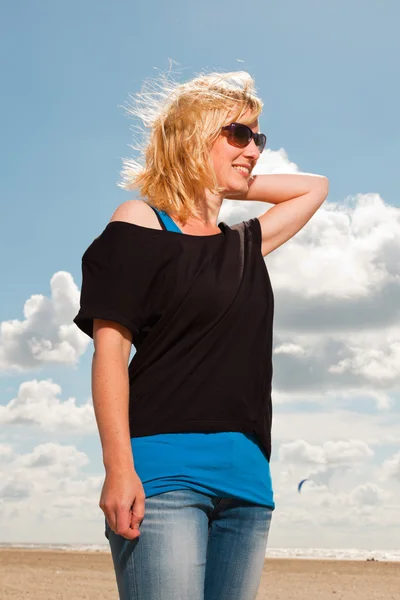 Gelukkig vrij blonde vrouw op het strand. genieten van de natuur. blauwe bewolkte hemel. zwarte trui en zonnebril dragen. — Stockfoto
