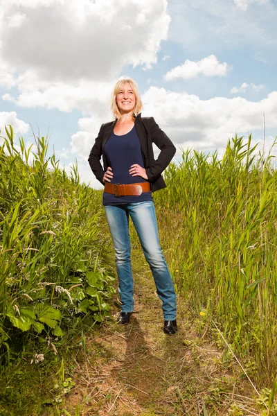 Glad ganska ung blond kvinna njuter av naturen. fält med högt gräs. blå mulen himmel. Svart jacka. — Stockfoto