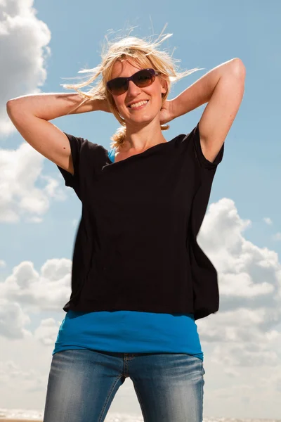 Mulher loira bonita feliz na praia. Desfrutando da natureza. Céu azul nublado. Vestindo camisola preta e óculos escuros . — Fotografia de Stock