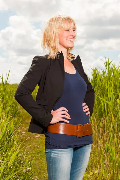 Lykkelig ung blond kvinne som nyter naturen. Felt med høyt gress. Blå skyet himmel. Svart jakke . – stockfoto