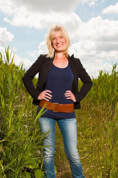 Zadowolony ładny blond kobieta z przyrodą. pole z wysokiej trawy. błękitne niebo pochmurne. Czarna kurtka. — Zdjęcie stockowe