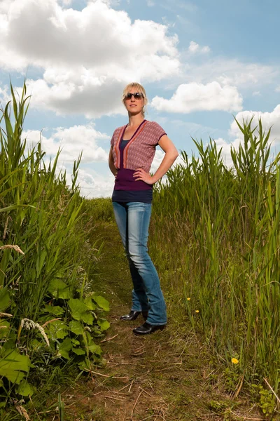 Fericit destul de tânără blondă cu ochelari de soare care se bucură de natură. Câmp cu iarbă înaltă. Albastru cer noros. Îmbrăcat în roșu . — Fotografie, imagine de stoc