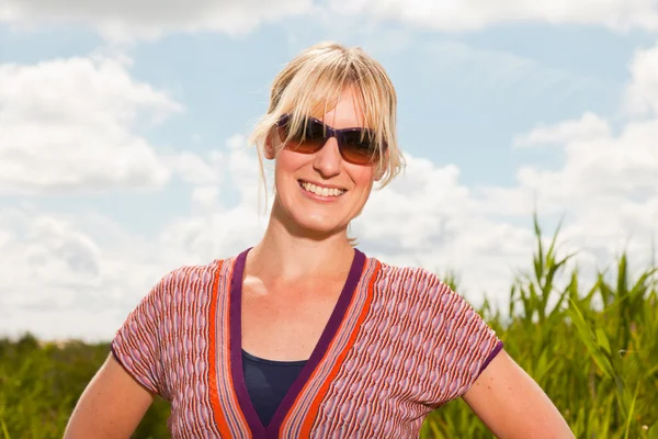 Felice bella giovane donna bionda con occhiali da sole godendo la natura. Campo con erba alta. Cielo blu nuvoloso. Vestita di rosso . — Foto Stock
