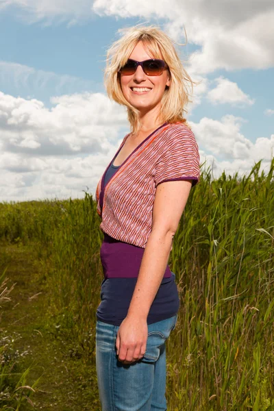 Happy pretty young blond woman with sunglasses enjoying nature. Field with high grass. Blue cloudy sky. Dressed in red. — Stock Photo, Image