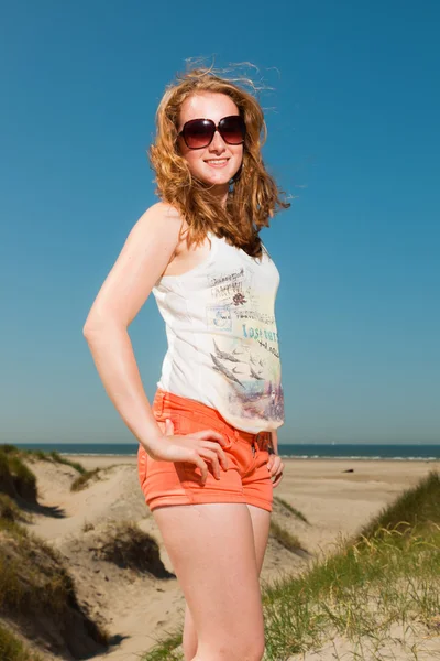 Chica bonita feliz con el pelo rojo largo y gafas de sol disfrutando de dunas de arena cerca de la playa en los veranos calurosos día. Cielo azul claro . —  Fotos de Stock