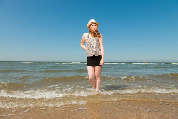 Glad söt flicka med långa röda hår och hatt njuta av uppfriskande stranden på varm sommardag. klar blå himmel. — Stockfoto
