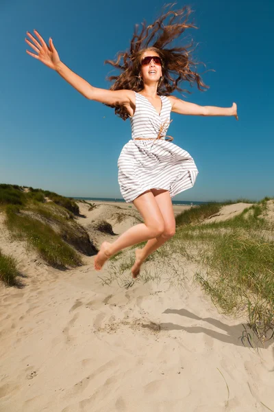 Happy pretty girl with long brown hair enjoying sand dunes near the beach on hot summers day. Wearing singlasses. Clear blue sky. — Stock Photo, Image