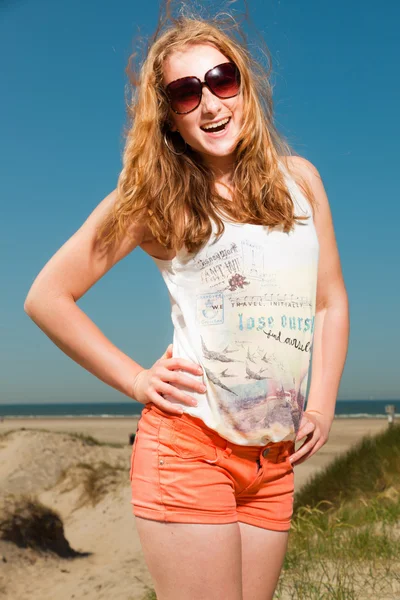 Happy pretty girl with long red hair and sunglasses enjoying sand dunes near the beach on hot summers day. Clear blue sky. — Stock Photo, Image