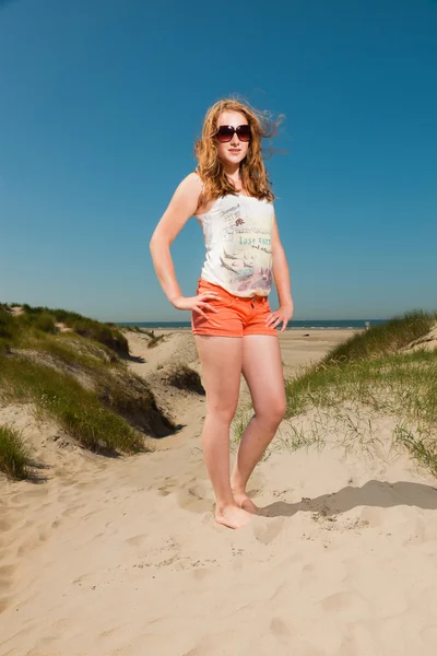 Jolie fille avec de longs cheveux roux et des lunettes de soleil profitant des dunes de sable près de la plage par une chaude journée d'été. Ciel bleu clair . — Photo