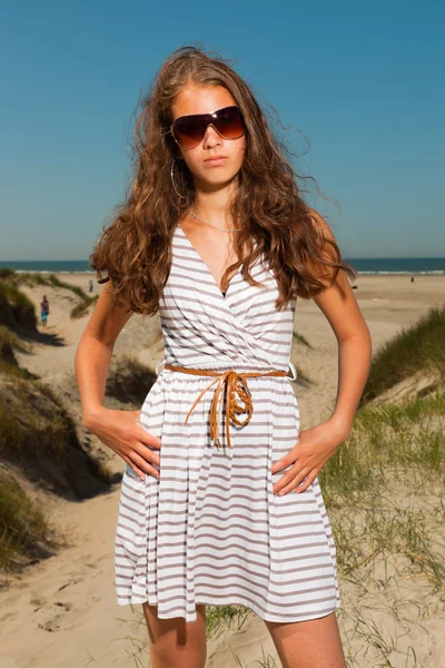 Happy pretty girl with long brown hair enjoying sand dunes near the beach on hot summers day. Wearing singlasses. Clear blue sky. — Stock Photo, Image