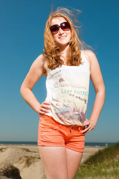 Ευτυχισμένο το όμορφο κορίτσι με μακριά κόκκινα μαλλιά και τα γυαλιά ηλίου απολαμβάνοντας αμμόλοφους, κοντά στην παραλία για την ημέρα καλοκαίρια. σαφή μπλε ουρανό. — Φωτογραφία Αρχείου