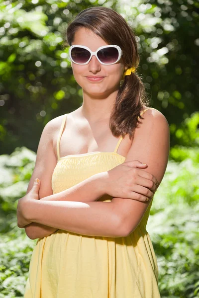 Mujer joven y bonita con cabello castaño largo disfrutando de la naturaleza en el bosque. Fondo verde borroso. Vestido amarillo y gafas de sol blancas . — Foto de Stock