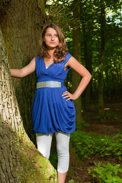 Bella giovane donna con lunghi capelli castani godersi la natura nella foresta. Sfondo fogliare verde. Indossa un vestito blu . — Foto Stock