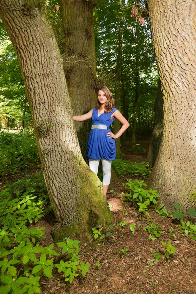 Pretty young woman with long brown hair enjoying nature in forest. Green foliage background. Wearing blue dress. — Stock Photo, Image