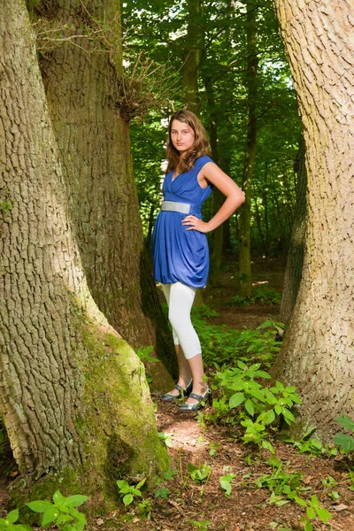 Pretty young woman with long brown hair enjoying nature in forest. Green foliage background. Wearing blue dress. — Stock Photo, Image