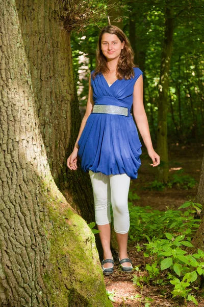 Досить молода жінка з довгим коричневим волоссям насолоджується природою в лісі. Зелене листя фону. Носіння синьої сукні . — стокове фото