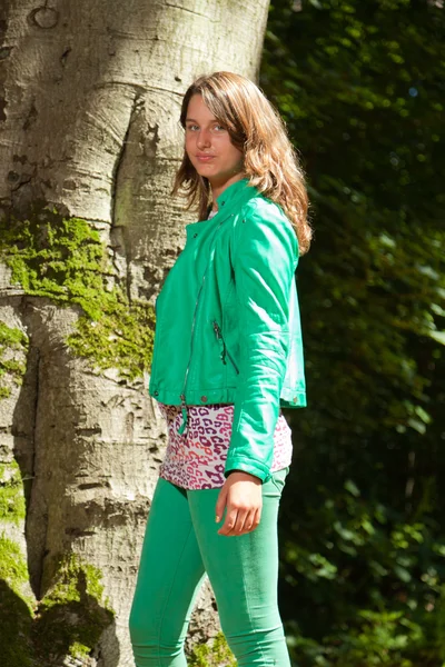 Pretty young woman with long brown hair enjoying nature in forest. Foliage in background. Wearing green jacket and trousers. Cool looking. Tough girl. — Stock Photo, Image