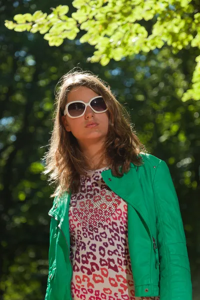 Mujer joven y bonita con cabello castaño largo disfrutando de la naturaleza en el bosque. Follaje en segundo plano. Usando chaqueta verde y pantalones y gafas de sol blancas. Bonito aspecto. Chica dura. . — Foto de Stock