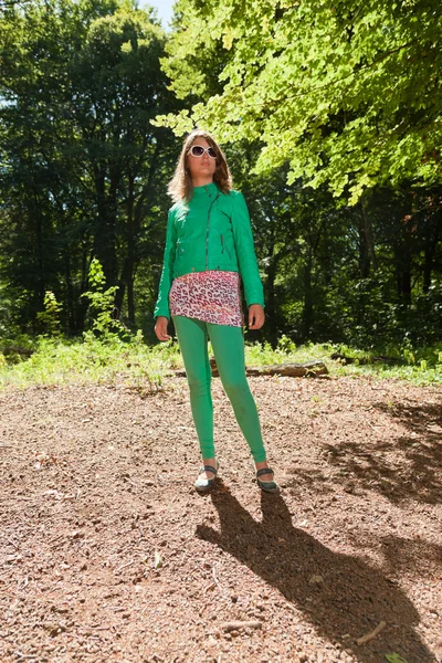 Pretty young woman with long brown hair enjoying nature in forest. Foliage in background. Wearing green jacket and trousers and white sunglasses. Cool looking. Tough girl. — Stock Photo, Image