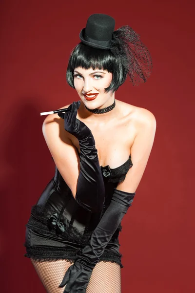 Burleske pin-up vrouw met zwarte haren in het zwart gekleed. sexy pose. roken sigaretten. het dragen van zwarte hoed. Studio mode schot geïsoleerd op rode achtergrond. — Stockfoto