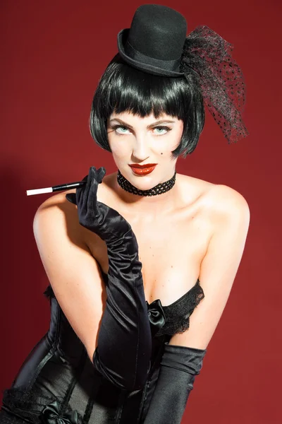 Burlesque pin up woman with black hair dressed in black. Sexy pose. Smoking cigarette. Wearing black hat. Studio fashion shot isolated on red background. — Stock Photo, Image