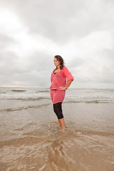 Pretty young woman enjoying outdoor nature near the beach. Standing in the water. Brown hair. Wearing pink shirt. Cloudy sky. — Stock Photo, Image