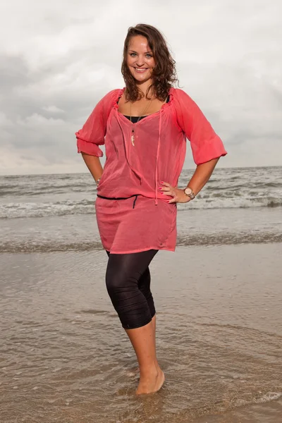 Mujer joven y feliz disfrutando de la naturaleza al aire libre cerca de la playa. De pie en el agua. Cabello castaño. Llevando camisa rosa. Cielo nublado . —  Fotos de Stock
