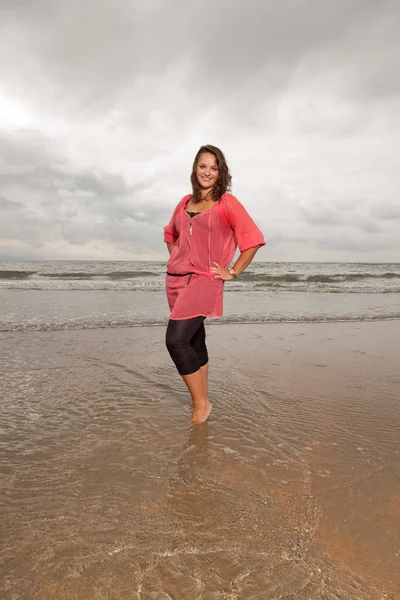 Щаслива молода жінка насолоджується природою на свіжому повітрі біля пляжу. Стоїть у воді. Коричневе волосся. На рожевій сорочці. Хмарне небо . — стокове фото