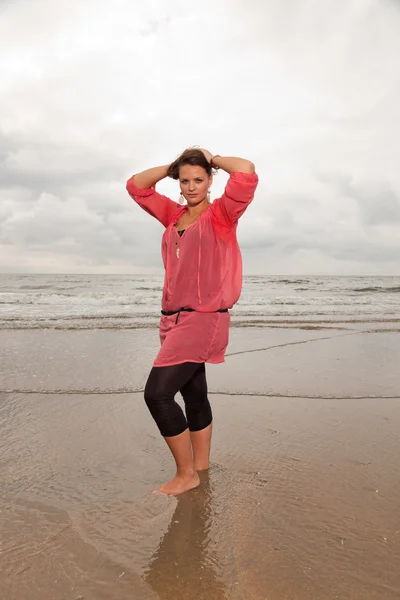 Happy young woman enjoying outdoor nature near the beach. Standing in the water. Brown hair. Wearing pink shirt. Cloudy sky. — Stock Photo, Image