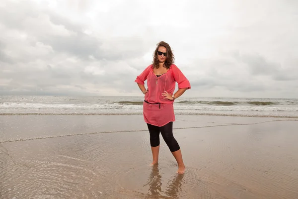 Mujer joven y feliz disfrutando de la naturaleza al aire libre cerca de la playa. De pie en el agua. Cabello castaño. Con camisa rosa y gafas de sol negras. Cielo nublado . —  Fotos de Stock