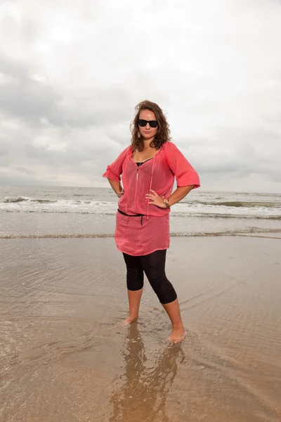 Happy young woman enjoying outdoor nature near the beach. Standing in the water. Brown hair. Wearing pink shirt and black sunglasses. Cloudy sky. — Stock Photo, Image