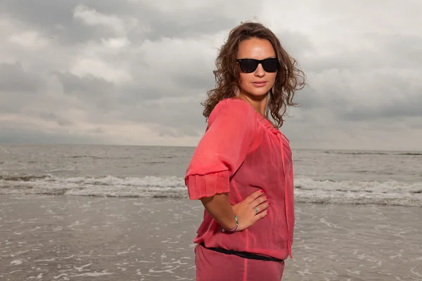Glückliche junge Frau, die die Natur in Strandnähe genießt. braune Haare. trägt rosa Hemd und schwarze Sonnenbrille. Bewölkter Himmel. — Stockfoto