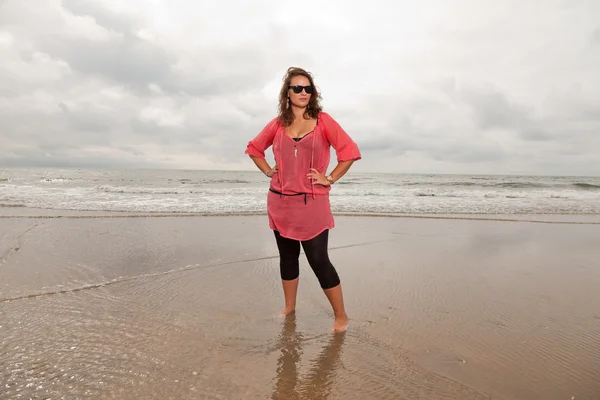Mujer joven y feliz disfrutando de la naturaleza al aire libre cerca de la playa. De pie en el agua. Cabello castaño. Con camisa rosa y gafas de sol negras. Cielo nublado . —  Fotos de Stock