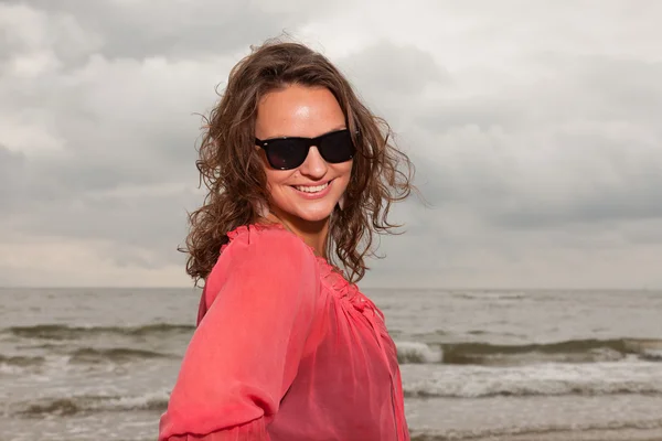 Joyeux jeune femme appréciant la nature en plein air près de la plage. Cheveux bruns. Porter une chemise rose et des lunettes de soleil noires. Ciel nuageux . — Photo