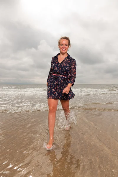 Csinos, fiatal nő élvezi a kültéri jellegét, a strand közelében. séta a vízben. vörös hajú. sötét kék ruhát visel. felhős ég. — Stock Fotó