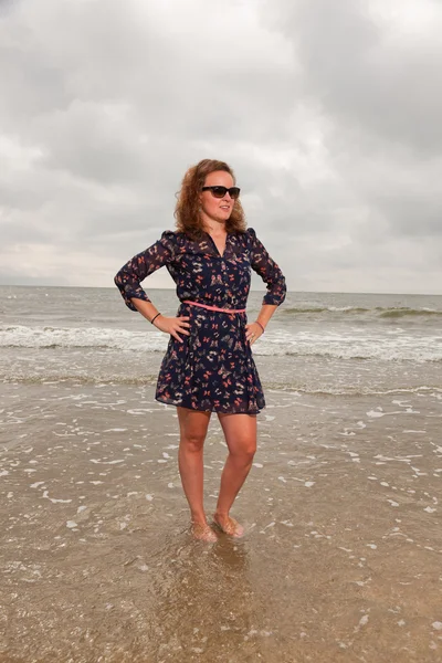 Pretty young woman enjoying outdoor nature near the beach. Standing in the water. Red hair. Wearing dark blue dress and black sunglasses. Cloudy sky. — Stock Photo, Image