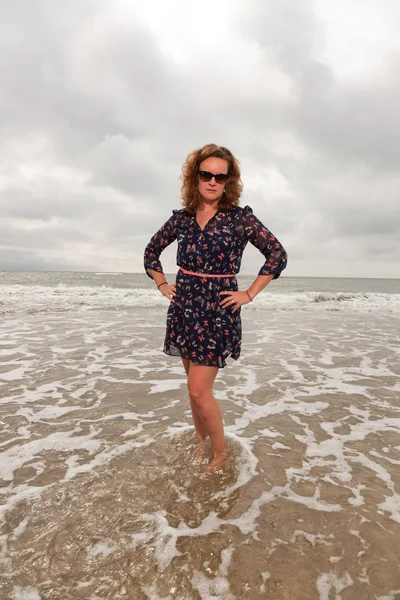 Bella giovane donna che si gode la natura all'aperto vicino alla spiaggia. In piedi in acqua. Capelli rossi. Indossa un vestito blu scuro e occhiali da sole neri. Cielo nuvoloso . — Foto Stock