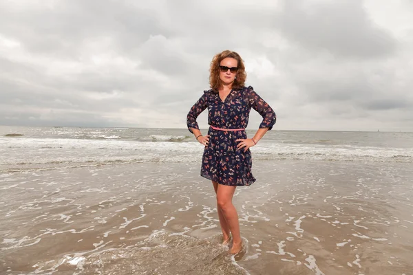 Hübsche junge Frau, die die Natur in Strandnähe genießt. im Wasser stehend. rote Haare. trägt ein dunkelblaues Kleid und eine schwarze Sonnenbrille. Bewölkter Himmel. — Stockfoto
