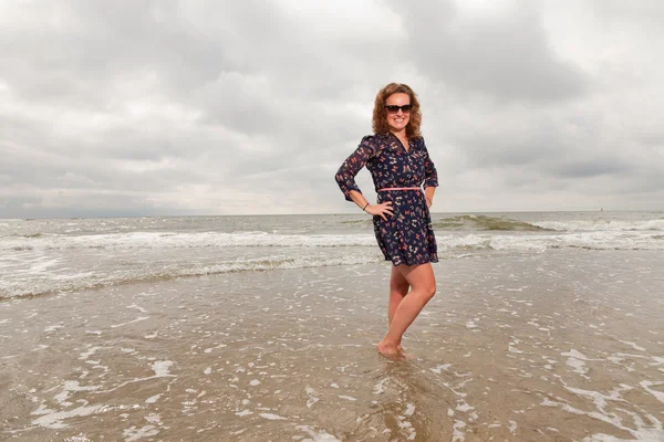 Ganska ung kvinna njuta utomhus natur nära stranden. stående i vattnet. rött hår. klädd i mörk blå klänning och svarta solglasögon. molnig himmel. — Stockfoto