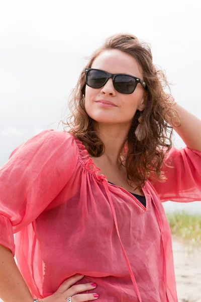 Mujer joven y feliz disfrutando de la naturaleza al aire libre cerca de la playa. Cabello castaño. Con camisa rosa y gafas de sol negras. Cielo nublado . — Foto de Stock