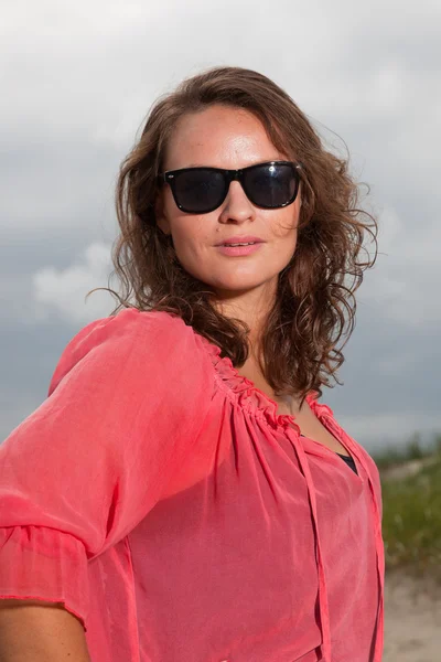 快乐的年轻女人享受海滩附近的户外自然。棕色的头发。身穿粉红色的衬衫和黑色太阳镜。多云的天空. — 图库照片