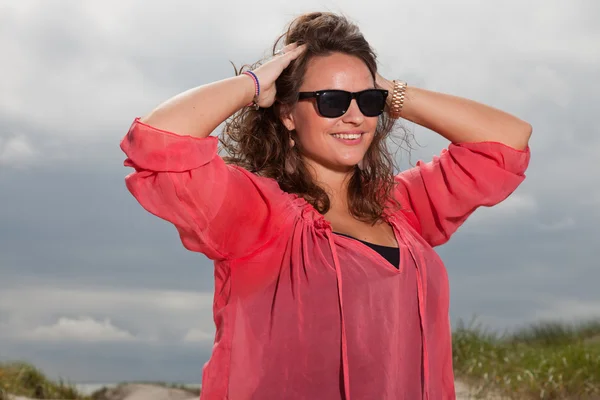 Joyeux jeune femme appréciant la nature en plein air près de la plage. Cheveux bruns. Porter une chemise rose et des lunettes de soleil noires. Ciel nuageux . — Photo