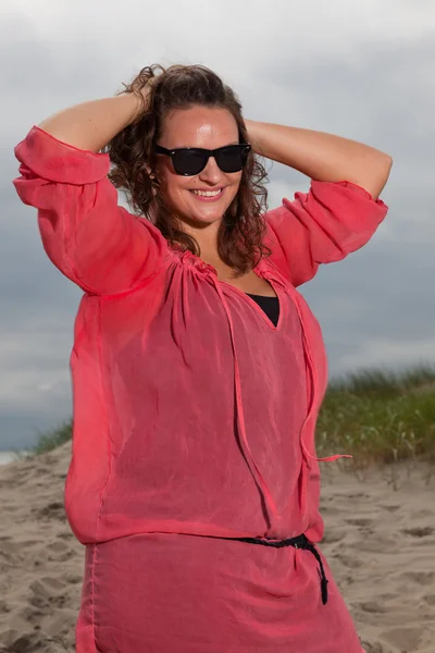 Щаслива молода жінка насолоджується природою на свіжому повітрі біля пляжу. Коричневе волосся. Носіння рожевої сорочки та чорних сонцезахисних окулярів. Хмарне небо . — стокове фото
