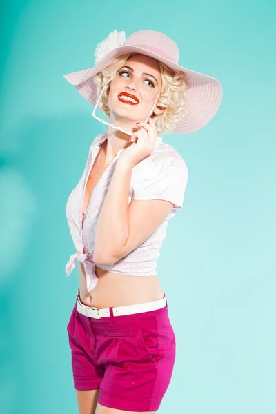 セクシーな金髪ガールアップピン帽子身に着けているピンクの t シャツとホットパンツ サングラスを保持しています。レトロなスタイル。ファッションはスタジオ撮影に分離の明るい青の背景. — ストック写真
