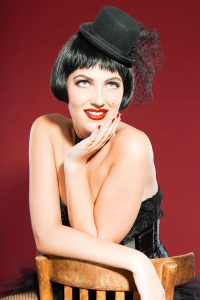 Burlesco pin up mulher com cabelo preto vestido de preto. Uma pose sexy. Sentado na cadeira. A usar chapéu preto. Estúdio moda tiro isolado no fundo vermelho . — Fotografia de Stock