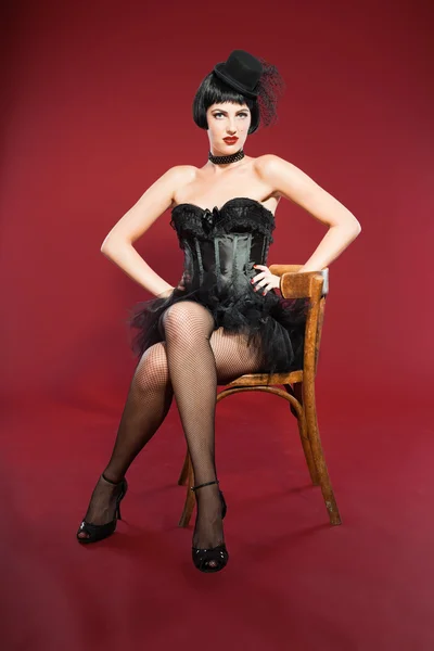 Burlesque pin up woman with black hair dressed in black. Sexy pose. Sitting on chair. Wearing black hat. Studio fashion shot isolated on red background. — Stock Photo, Image