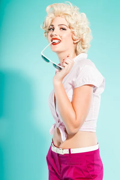 Sexy blonde pin-up meisje dragen roze shirt en hotpants houden zonnebril. retro stijl. mode studio opname geïsoleerd op lichte blauwe achtergrond. — Stockfoto