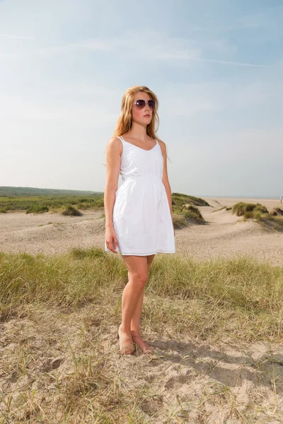 Hezká dívka s červenými dlouhé vlasy nosí bílé šaty a sluneční brýle, užívat přírodu v blízkosti pláže. horký letní den s modré oblohy jasno. — Stock fotografie