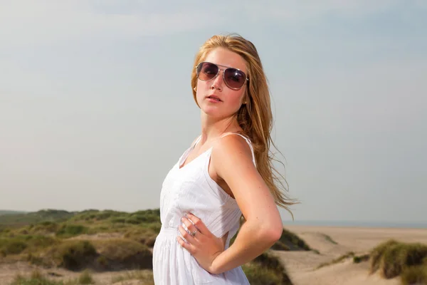 Bella ragazza con i capelli lunghi rossi indossa abito bianco e occhiali da sole godendo la natura vicino alla spiaggia. Caldo giorno d'estate con cielo nuvoloso blu . — Foto Stock
