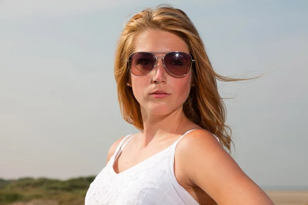 Söt flicka med rött långt hår, klädd i vit klänning och solglasögon som njuter av naturen nära stranden. varm sommardag med blå mulen himmel. — Stockfoto
